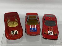 Three red vintage Hot Wheels cars displayed side by side on a clear riser, showing front and top details for each car.