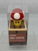 Front view of wine stopper in sealed transparent box with red toadstool mushroom top and brown block base with text.