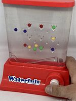 Front view of Waterfuls toy game showing colored balls inside water chamber and white button on red base.
