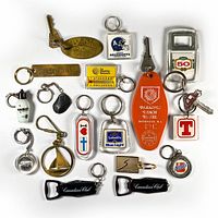 Top view of all 17 vintage keychains arranged on white background, showing variety of materials and brands including brass, plastic, and metal pieces.