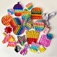 Overview of 23 colorful Pop It fidget toys laid out on white surface, shapes include food items, hearts, circles, star, unicorn, strawberry clutch, and multiple keychain-sized toys.