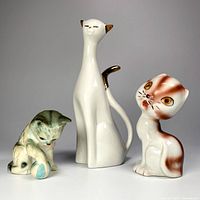 Three vintage ceramic cat figurines displayed together including Lefton kitten, tall unmarked cat, and big-headed cat marked Japan.