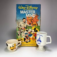 Front view of the Walt Disney Master Mind game box along with the two mugs: a milk glass blue-and-white Joan mug and a small cream-colored studio pottery character mug with Disney faces.