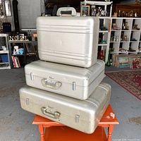 Three vintage Halliburton aluminum suitcases stacked on an orange table. The suitcases are silver in color with some surface wear. Visible metal latches and molded handles on each.