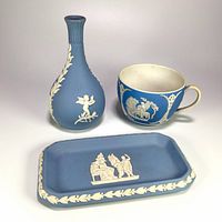 Photo showing all three Wedgwood jasperware pieces: bud vase, teacup, and rectangular pin tray, each decorated with white neoclassical relief scenes on pale blue backgrounds.