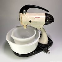 Front side view of Sunbeam Mixmaster with two milk glass mixing bowls on black base, cord wrapped around base, showing vintage wear.