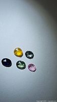 Five small sapphire gemstones laid out on white surface, showing mixed colors and rose cut style.