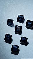 Seven natural princess cut blue sapphires with deep blue color and transparent quality, arranged on white surface.