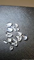 Mixed clear natural crystal quartz cabochon gemstones arranged on a dark textured surface, showing varied shapes and sizes
