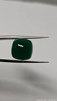 Photo showing an 8.36 carat natural vivid green Onyx cabochon gemstone held by tweezers, highlighting its cushion shape and glossy surface.