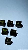 Six natural green princess cut tourmaline gemstones arranged together on a light blue background, showing square shape and clarity under lighting.
