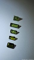 Photo showing five natural green tourmaline gemstones in emerald and baguette cuts arranged vertically on a white background. Colors range from light to medium green, clarity appears good.