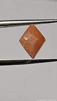 Close-up image of a natural peach moonstone fancy cut gemstone held by tweezers. Shows the kite-shaped cut, peach color, and internal sparkles.