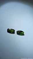 Two natural green chrome diopside gemstones laid flat on a white surface, one marquis cut and one cushion cut, showing deep green color and facets.
