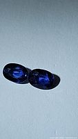Two oval cut royal blue sapphire gems placed side by side on a white background, showing the deep blue color and facets of the stones.