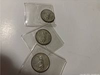 Three silver quarters with different centennial designs in individual protective sleeves on white surface