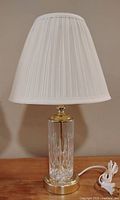 Single end table lamp with cream pleated fabric shade, gold-tone metal fittings, and clear cut glass cylindrical base with gold-tone metal round foot. Electrical cord and plug attached.