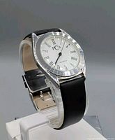 Side angle showing the watch face and black leather strap on a clear display stand.