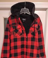 Red and black checkered men's hoodie hanging on a clear plastic hanger showing front view with chest pockets and hood lining.