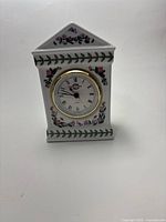 Front view of the white ceramic carriage clock showing the clock face with Roman numerals and surrounding floral decoration.