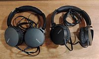 Two pairs of black Sony wired over-ear headphones with adjustable headbands and padded ear cups laid on a wooden surface.