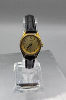 Front view of women's Cardinal wristwatch with gold-toned round case, ivory dial with black Arabic numerals, and black leather crocodile pattern strap, displayed on clear stand.