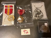 Two military medals, silver-tone necklace pendant dated 1983, and silver coin in plastic case all shown on a dark surface.