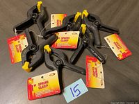 Top view of six new KC spring clamps with black bodies and yellow grips, tags attached, laid out on a dark surface.