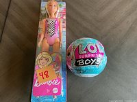 Barbie doll in pink swimsuit and L.O.L. Surprise Boys ball seen in original packaging against a neutral background.