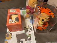 Photo showing two plush toys (Winnie the Pooh and Tigger) in clear display boxes, an orange box of paper cups with puppy faces, and a puppy-themed paper tablecloth beneath.