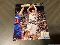 Signed basketball action photo of player #26 in Sixers jersey shooting with defender in blue jersey number 20 attempting to block.