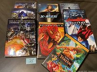 Photo displaying a collection of nine used DVDs with visible movie titles and artwork including Transformers, X-Men, Tarzan, and several superhero movies.