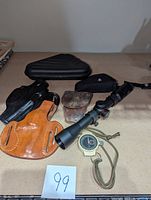 Photo of assorted holsters, handgun case, riflescope and stopwatch on beige surface.