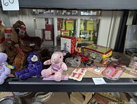 Shelf display of assorted plush bears including purple, brown, and pink embroidered bears, plus Rudolph The Red-Nosed Reindeer stuffed animals and other figurines and items.