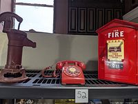 Three items on a metal rack: a red antique rotary phone, a red fire phone box marked Randix with emergency instructions, and a red metal hand pump.