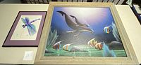 Both framed artworks displayed on a table side-by-side: larger underwater airbrush scene on right, smaller dragonfly watercolor with purple mat on left.