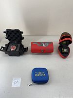 Five items arranged: black hardshell case with red dial watch, red embroidered cobra case cylinder, red helmet-style case with rectangular watch, blue zippered kit.