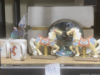 Front view of two ceramic carousel horse figurines and two mugs with carousel horse images, along with a decorative plate placed behind them.
