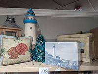 View of nautical themed items on shelf including lighthouse statues, seashell throw pillow, ceramic fish candle holder, wooden chest, and candle lantern.