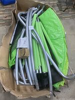 Box containing unassembled metal frame tubes with green padding and trampoline mat visible inside.