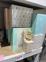 Stacked Precious Moments boxes in white, teal, and beige colors with porcelain figurines placed on top and in front of boxes.