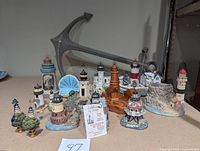 Photo showing assorted hand-painted resin and ceramic lighthouse figurines arranged in front of a large metal anchor wall decor. Includes ceramic clam shell tealight holder and various lighthouse bases.