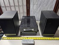 Full view of Sony stereo system main unit flanked by two speakers, with a white remote control in the foreground and measuring tape showing scale.