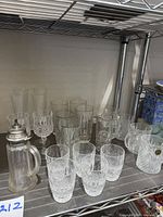 Overview of assorted crystal glasses including Waterford tumblers, wine glasses, Paul Sebastian mugs, and syrup pitcher placed on metal shelf.