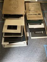 Two stacks of assorted mounting mats placed on carpeted floor. Mats vary in size and color, primarily black, white, cream, and dark green. Some mats partially covered by cardboard packaging showing minor wear.