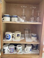 Photo of cupboard shelf showcasing three white mugs with blue stripes, three clear stemmed wine glasses, four graphic printed mugs, and one clear tumbler glass.