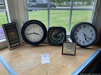 Five clocks and thermometers/wind gauge displayed on a wooden surface in front of a window, showing a mix of analog and digital devices.
