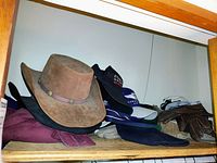 Shelf with brown Bailey suede cowboy hat, several baseball caps with embroidered logos, brown leather gloves and folded clothing items