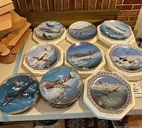 Overview of 10 collector plates depicting different WWII military aircraft arranged on a table, showing variety of round and octagonal shapes.
