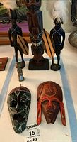 Photo of five carved wooden tribal art pieces including two warrior figurines with shields and feathers, a central totem pole statue, and two flat masks on a cloth surface.